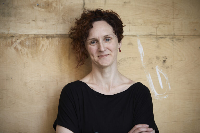 A colour head and shoulders photo of Mel Lambert with arms crossed and head tilted, smiling and wearing a black t-shirt