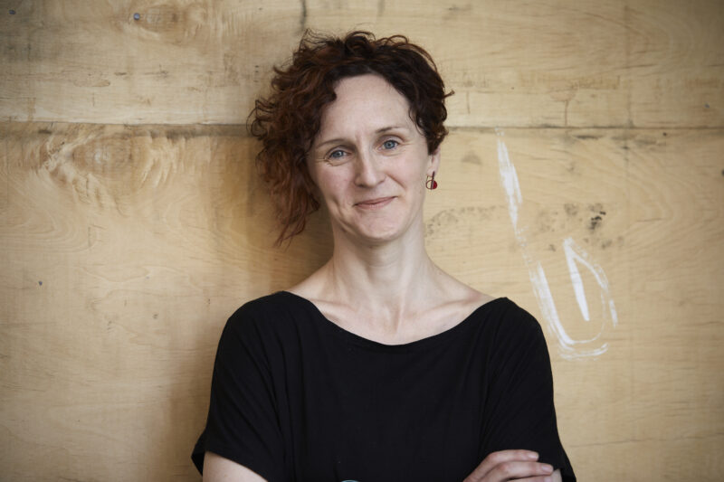 A colour head and shoulders photo of Mel Lambert with arms crossed and head tilted, smiling and wearing a black t-shirt