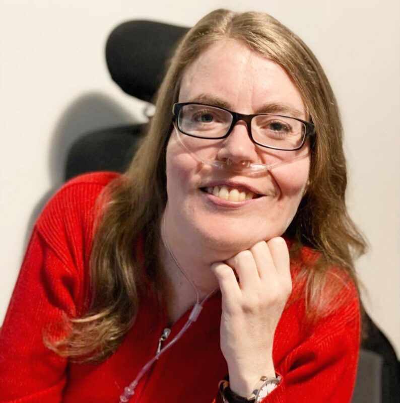 A colour head shot of Kelly McLean, wearing a red jacket and smiling directly into the camera