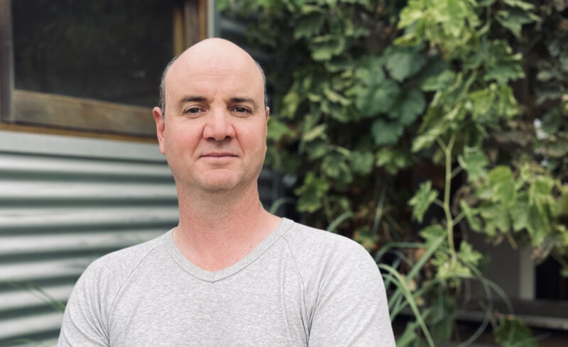 A white man in a grey t-shirt standing in front of a tree with green leaves and a building with silver corrugated wall.