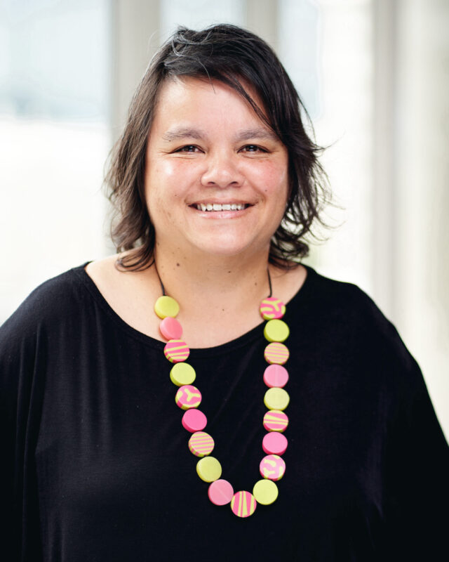 A picture of a woman with mid-length black hair smiling at the camera and wearing black top and pink, yellow and orange beads.