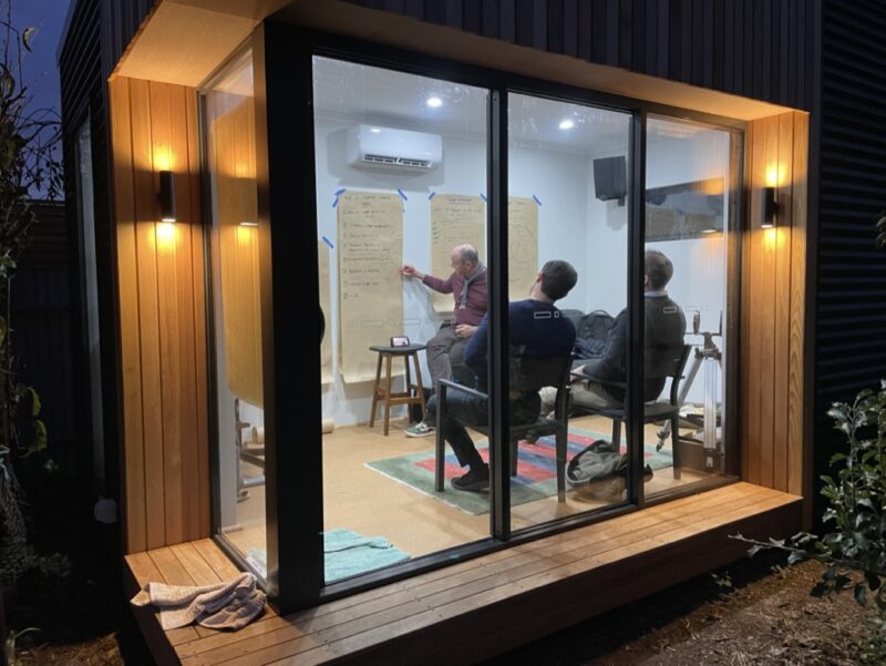 three men sit in a small, wood-clad office at night, with lights on inside. They are looking at a poster on the wall.