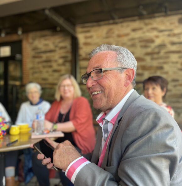 a man with glasses and grey hair, wearing a pink and white striped shirt and grey jacket. He is holding a phone and smiling, with other people in the background looking towards him.