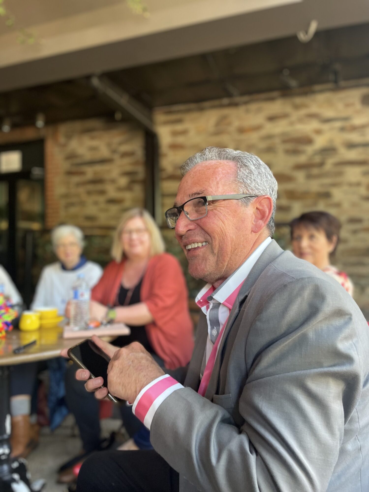 a man with glasses and grey hair, wearing a pink and white striped shirt and grey jacket. He is holding a phone and smiling, with other people in the background looking towards him.