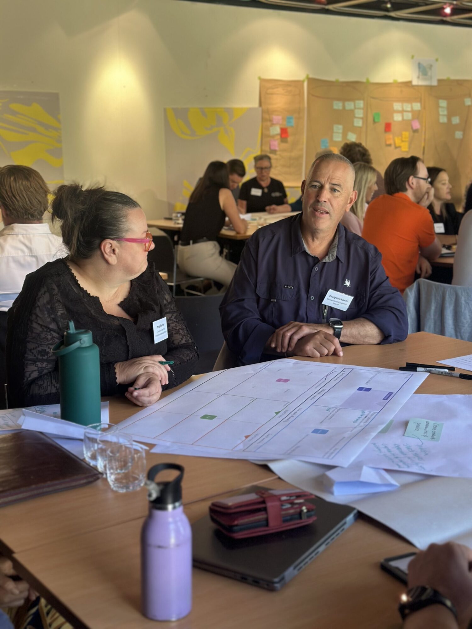 a man and a woman sit at a table in discussion with worksheets in front of them. Others in the background are also discussing at tables. 