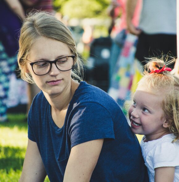a little girl with blond hair and cheeky grin sitting behind her mum, who looks back at her. They are in a sunny park.