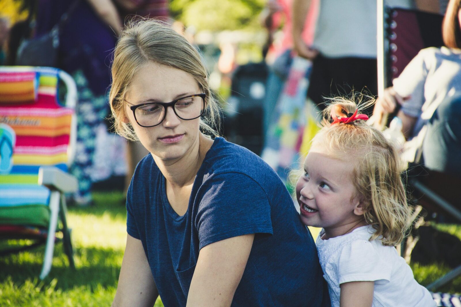 a little girl with blond hair and cheeky grin sitting behind her mum, who looks back at her. They are in a sunny park.