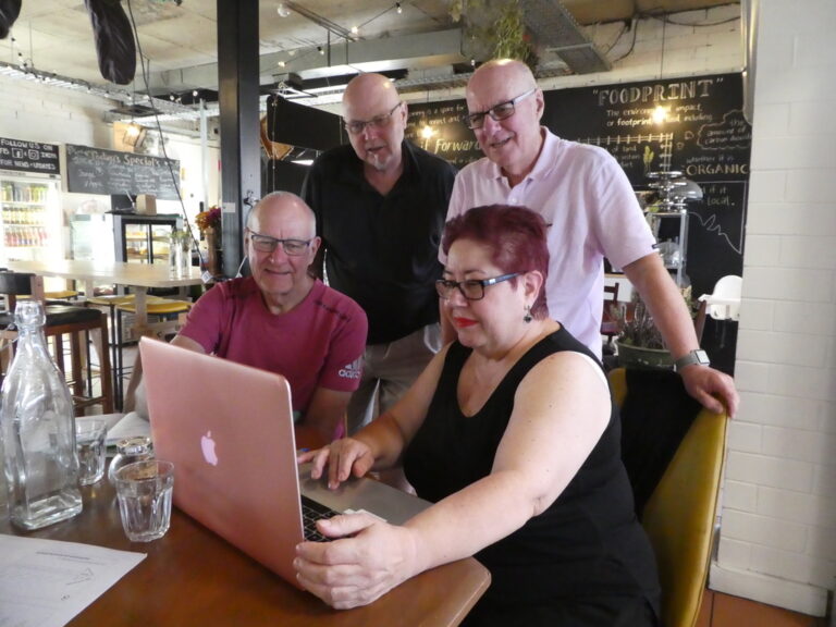 three men and a woman casually dressed in a cafe gathered around a computer looking at the screen.