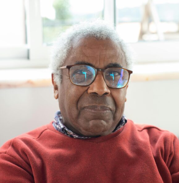 a black man with silver curly hair and glasses looks directly at the camera. He is sitting by a window wearing a red jumper.