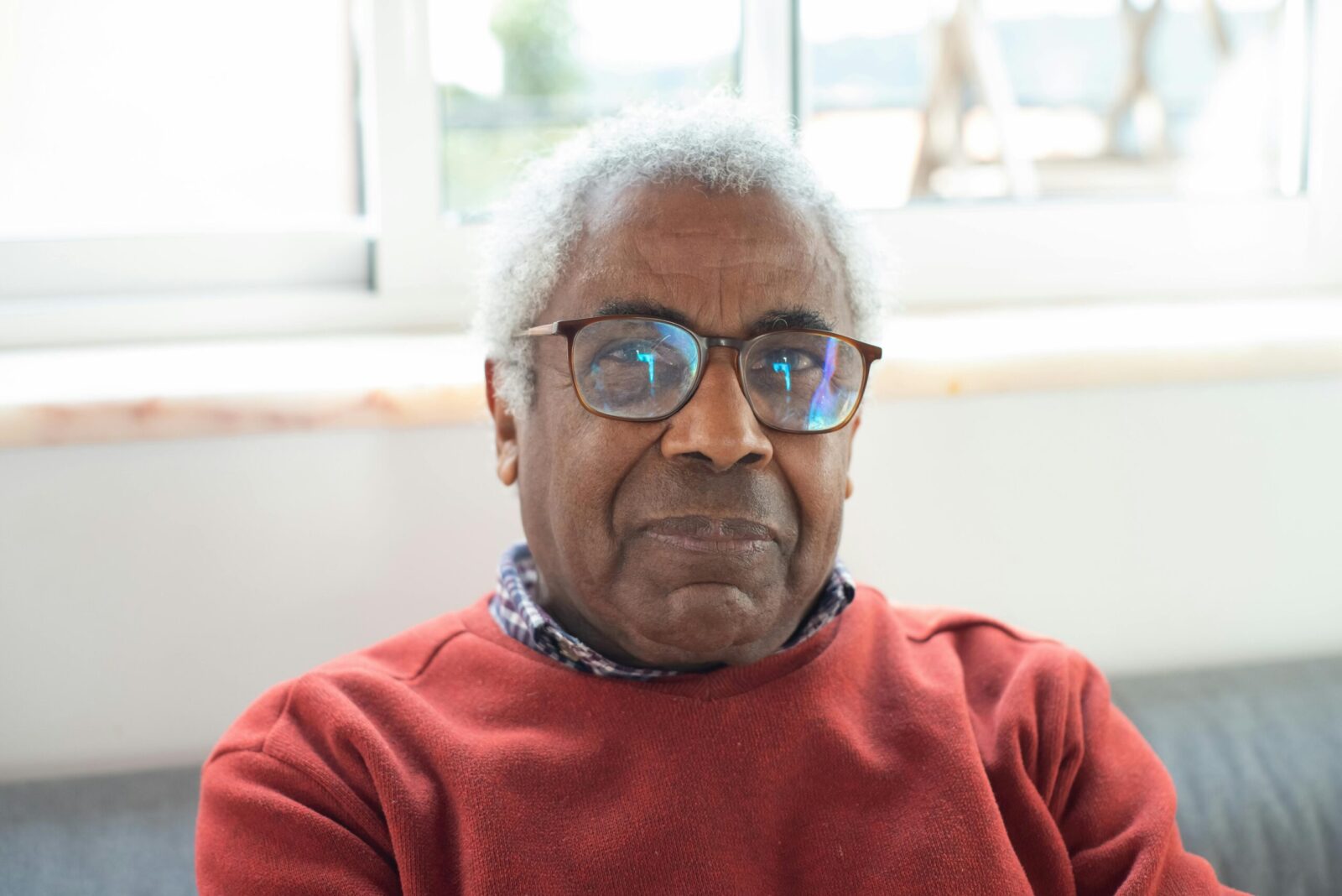 a black man with silver curly hair and glasses looks directly at the camera. He is sitting by a window wearing a red jumper.