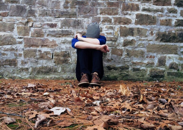 person sits on the ground amongst dried leaves and against a stone wall. They have knees pulled up and head buried behind their arms.