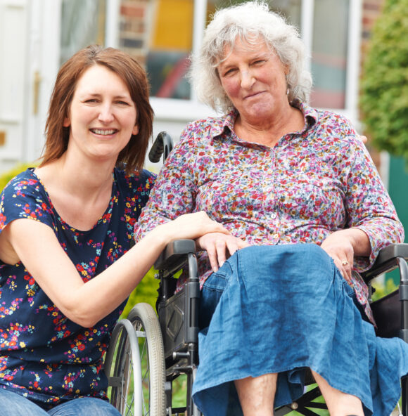 Two women smile at the camera. One woman has silver curly hair and is in a wheelchair, wearing a floral shirt; the other with long brown hair kneels beside her